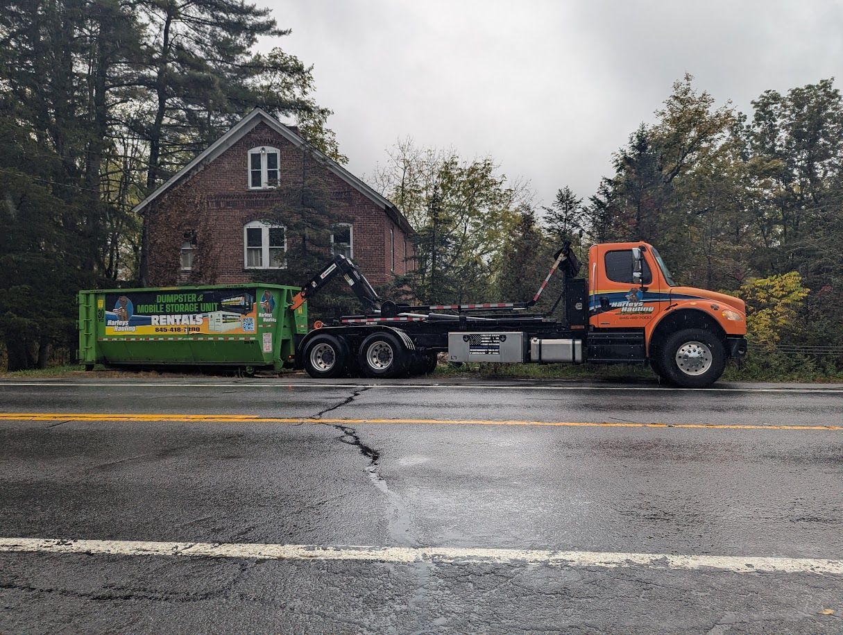 Orange semi-truck hauling a green dumpster trailer beside a house on a wet roadside