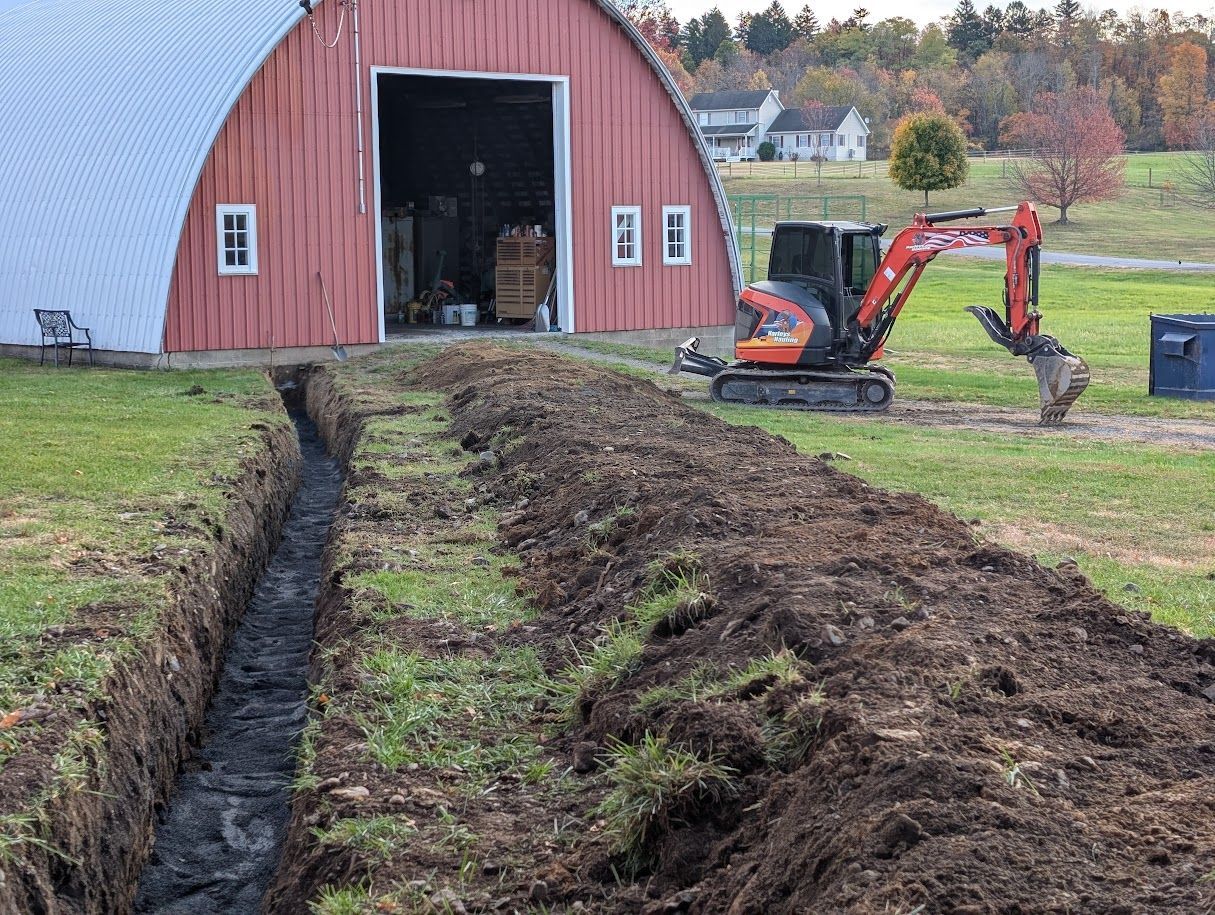 Mini excavator digging a trench beside a red barn in a grassy field