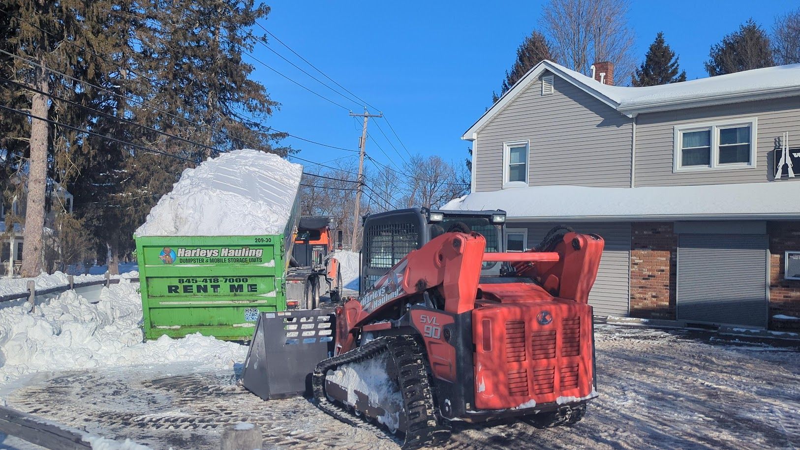 Red snowblower clearing a snowy driveway beside a green dumpster and a house in winter.