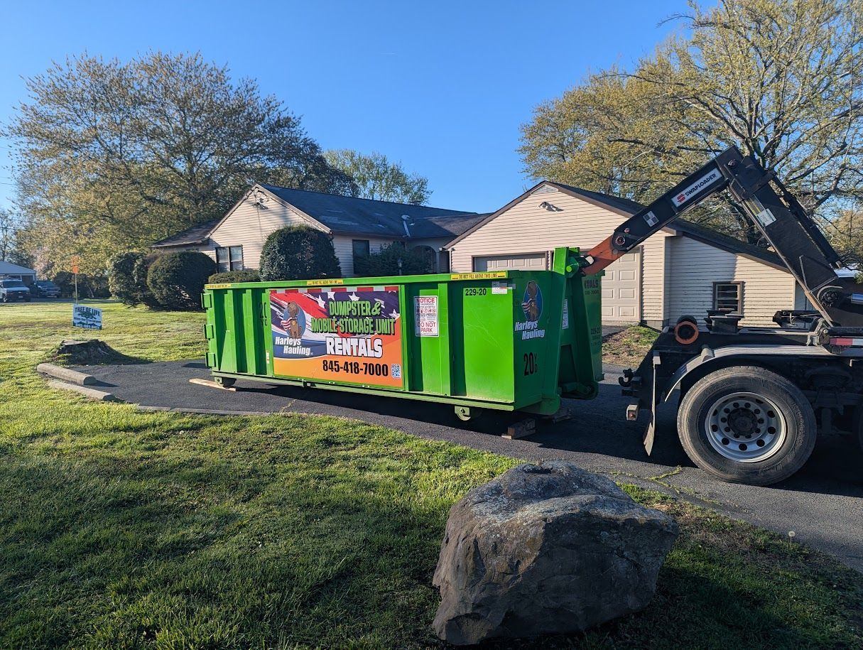 Green dumpster trailer beside a house in a sunny yard, with trees and a large rock in front.