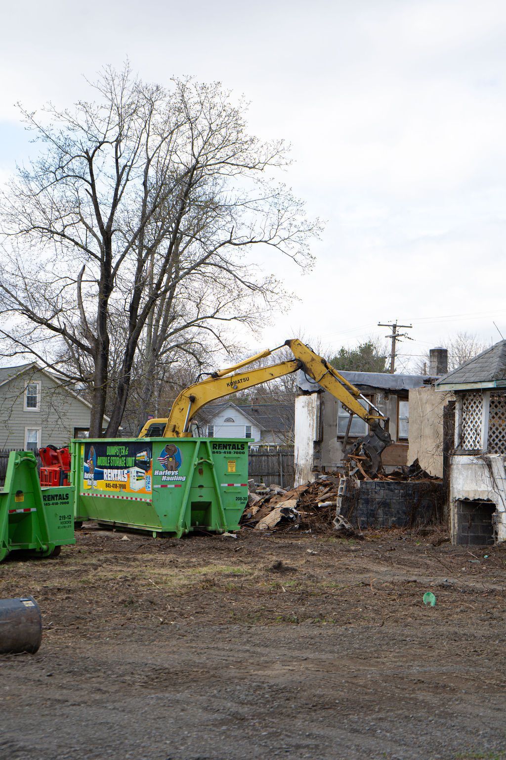 Excavator demolishing a damaged house beside a green dumpster and bare trees