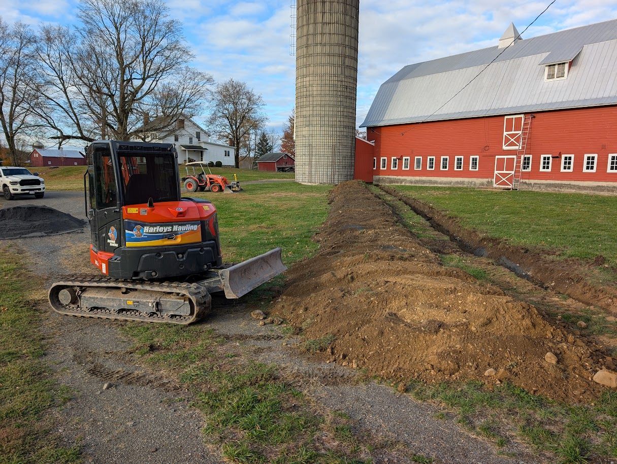 Small excavator leveling dirt beside a red barn and silo on a farm lane.