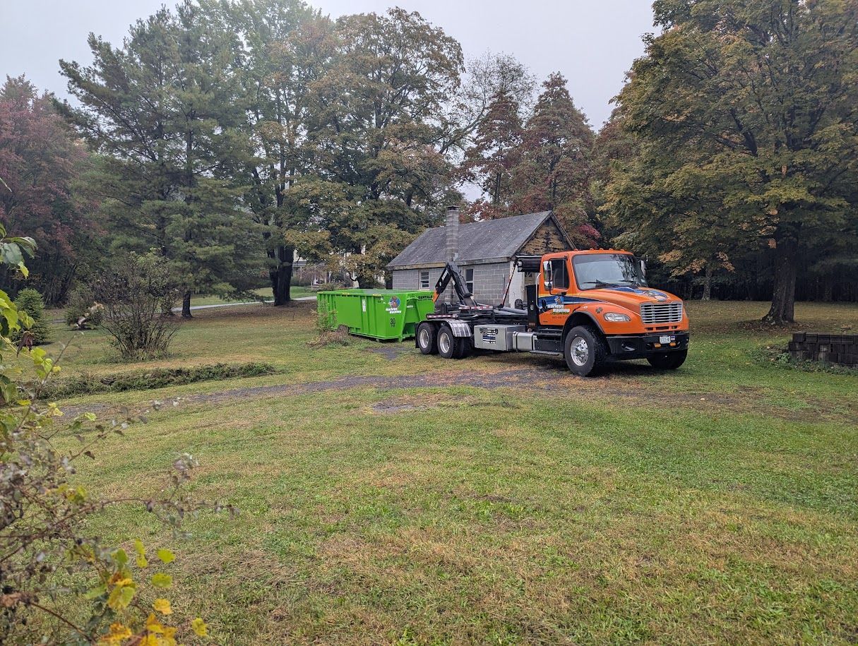 Orange truck towing green dumpster trailer in a grassy yard with trees and a house in the background