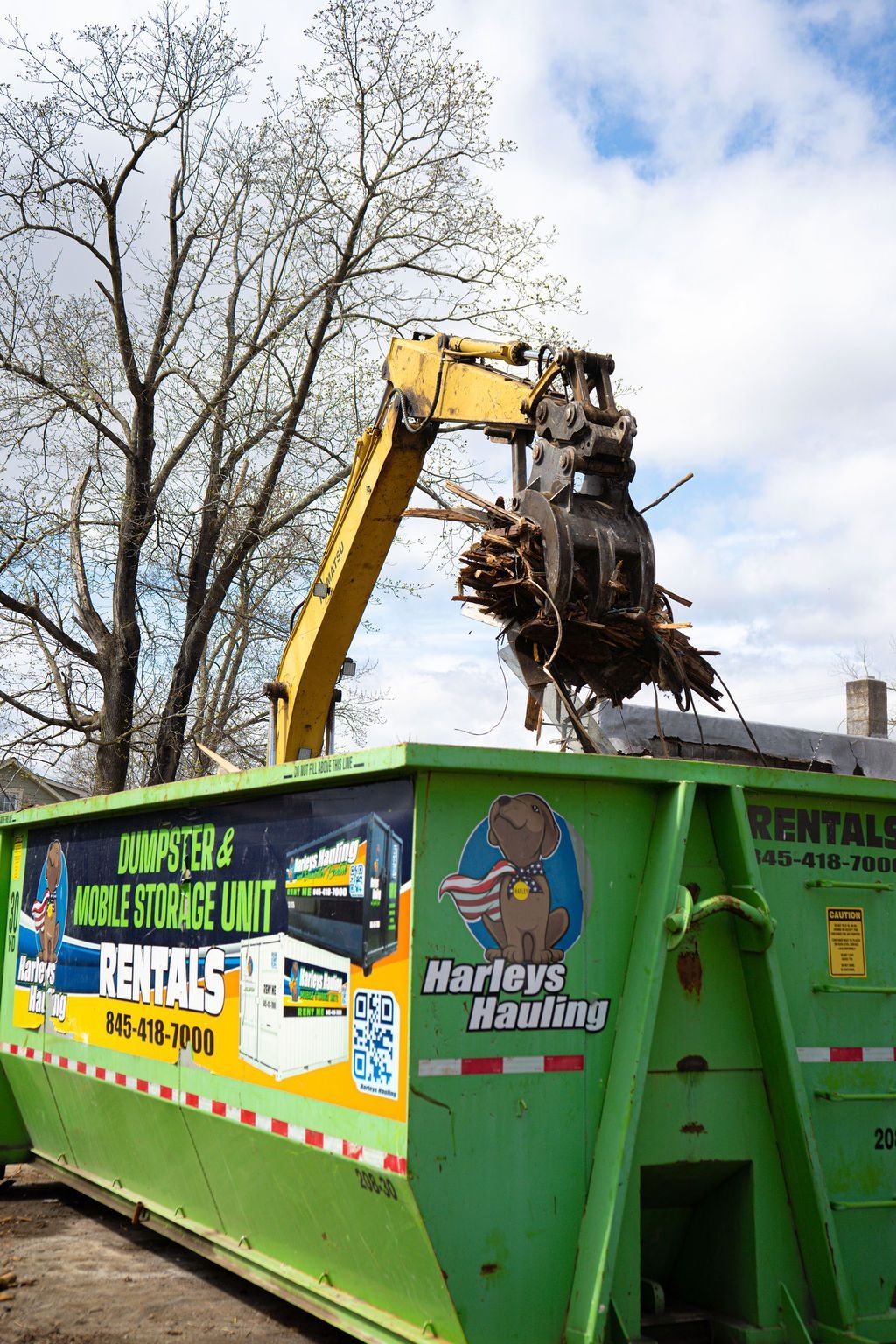 Yellow excavator loads debris into a green rental dumpster outdoors under a cloudy sky.