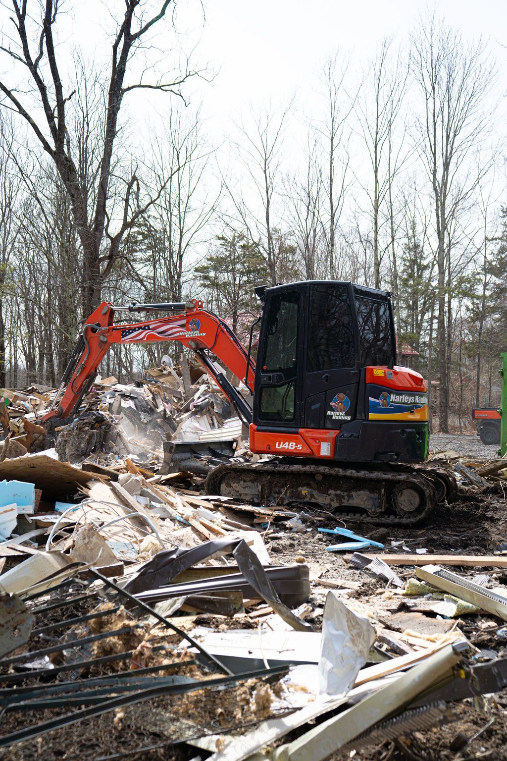 Orange excavator clearing debris in a wooded area with damaged building materials scattered around