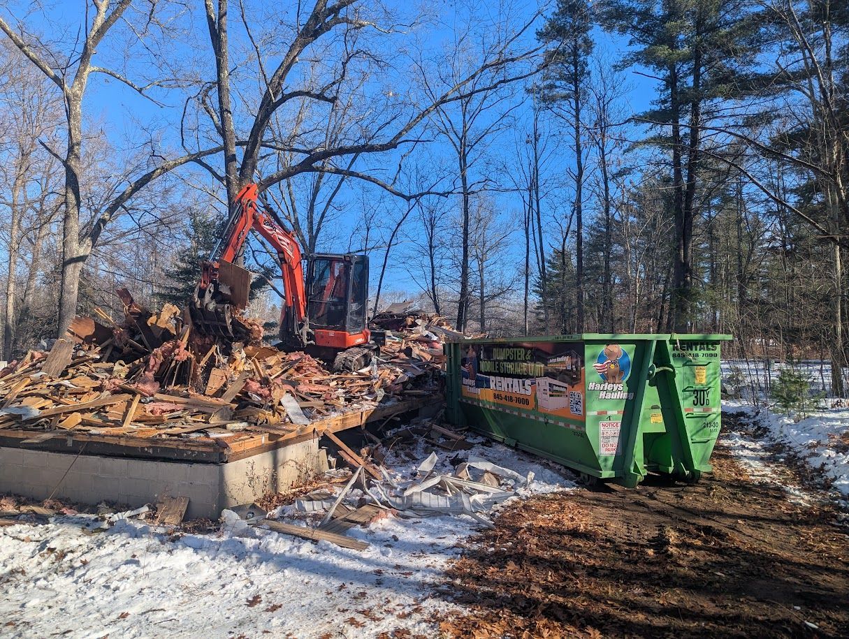 Excavator clearing rubble at a burned site beside a green dumpster in a snowy wooded area.
