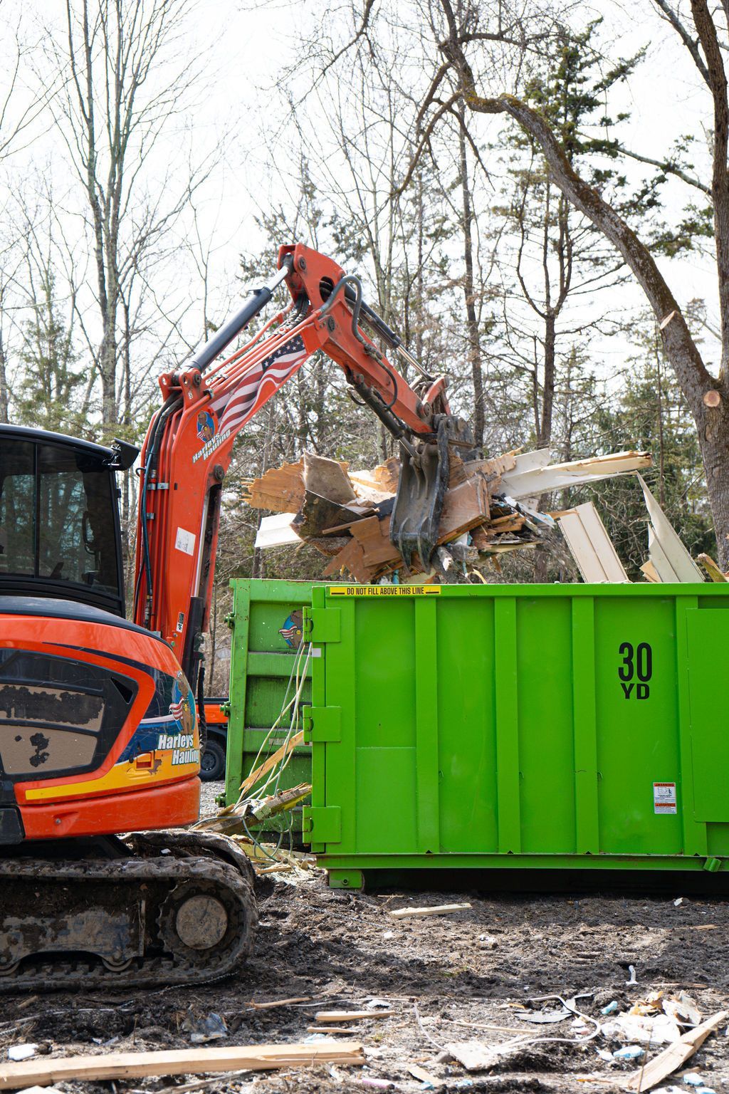 Orange excavator loading debris into a green dumpster at a wooded construction site