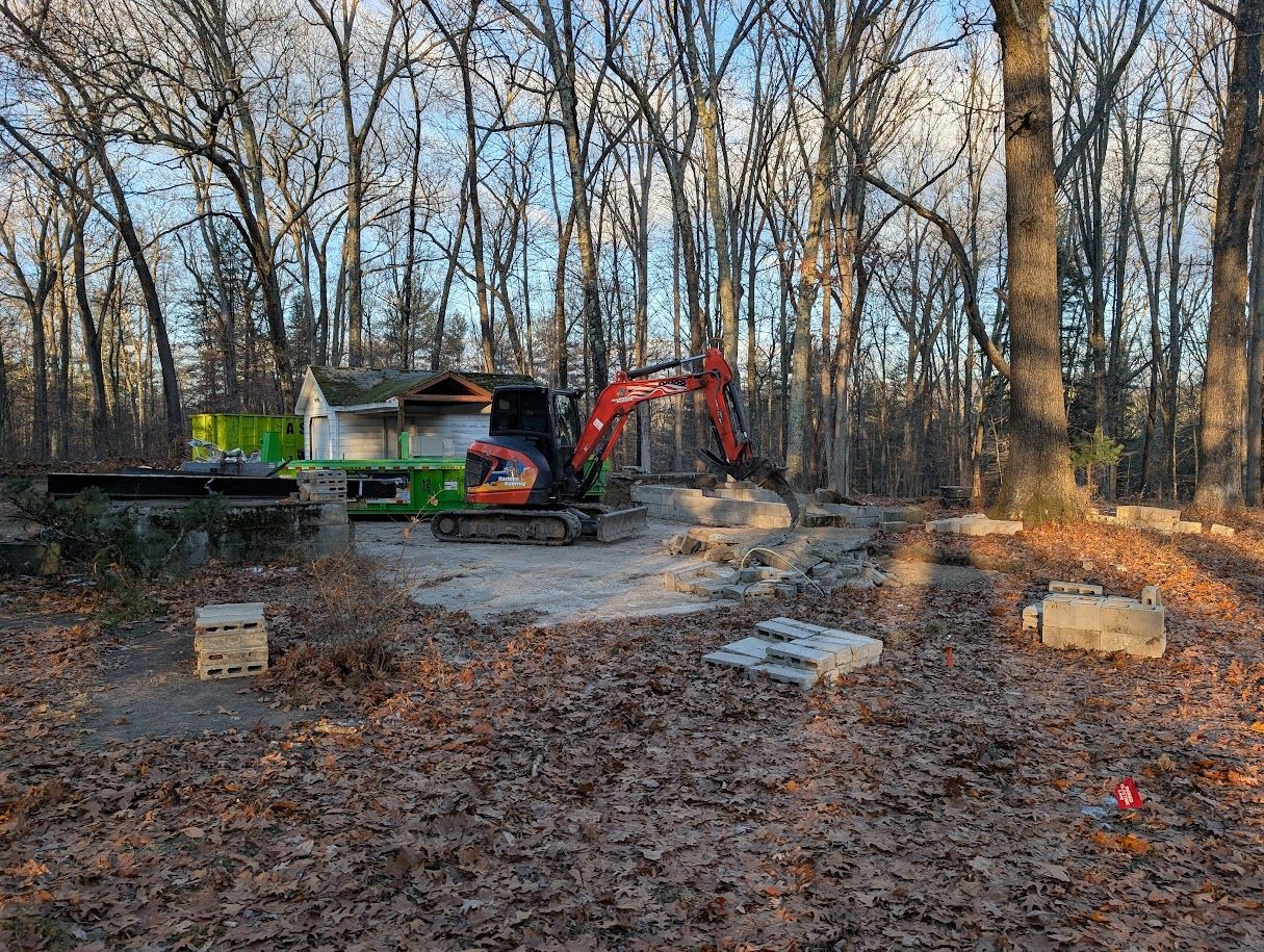 Excavator and construction equipment in a wooded clearing with bare trees and leaf-covered ground