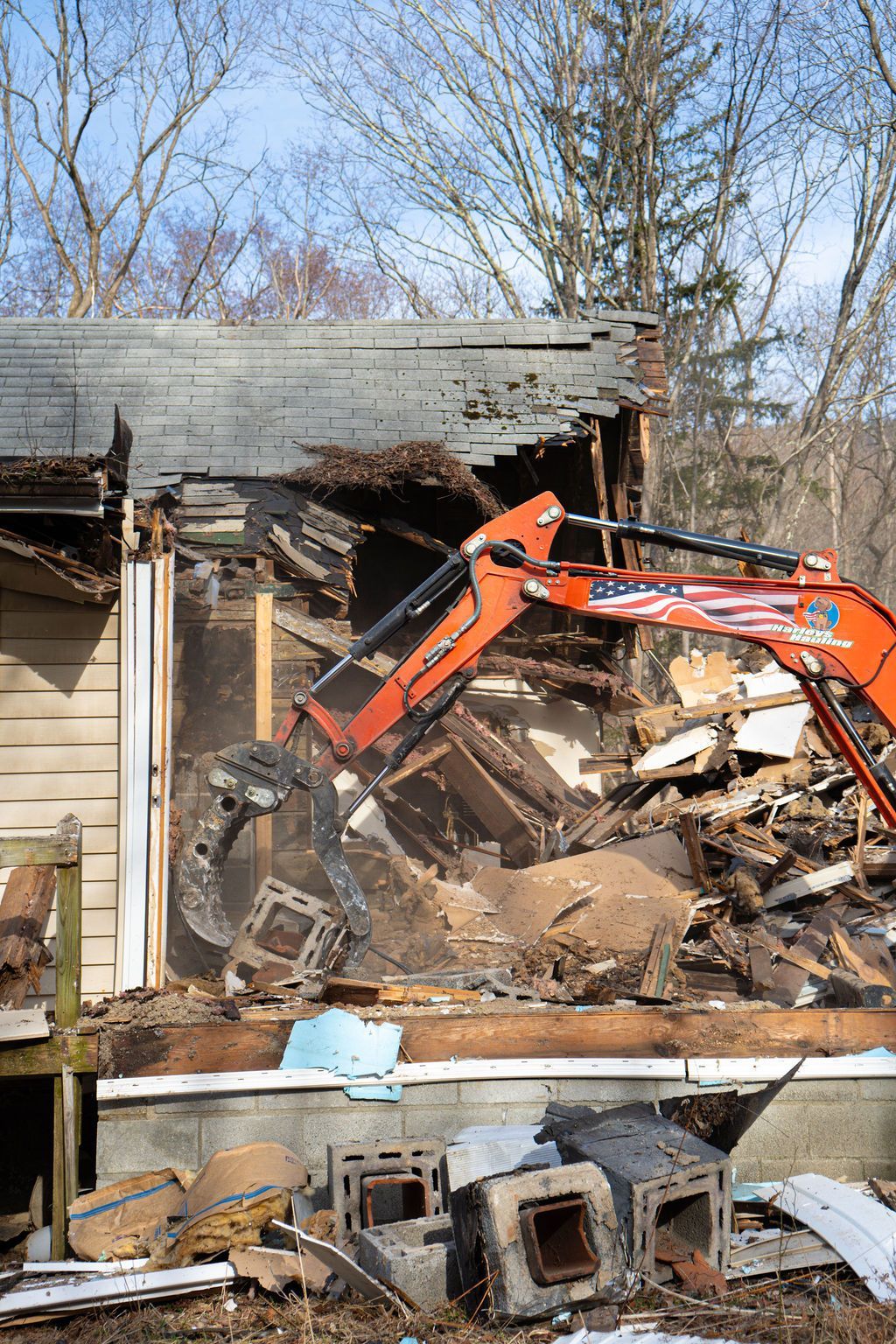 Orange excavator demolishing a damaged house amid debris and broken walls