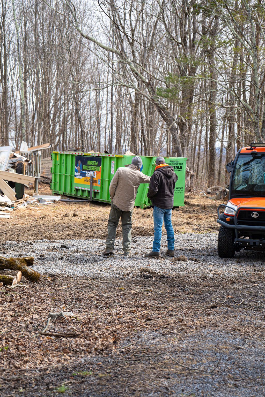 Two people standing by a green dumpster and orange truck in a wooded worksite