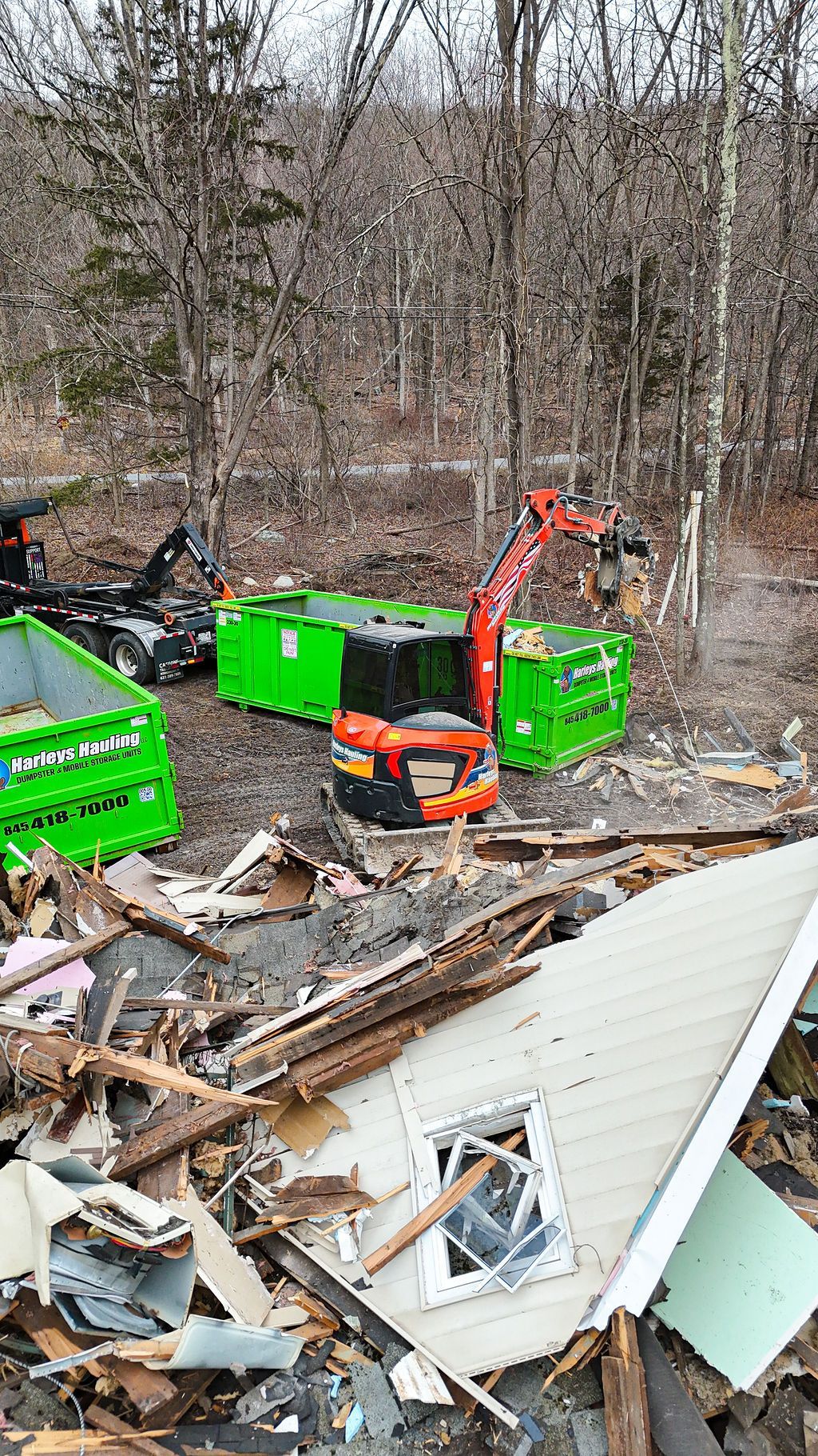 Demolition debris and an excavator beside bright green dumpsters in a wooded lot