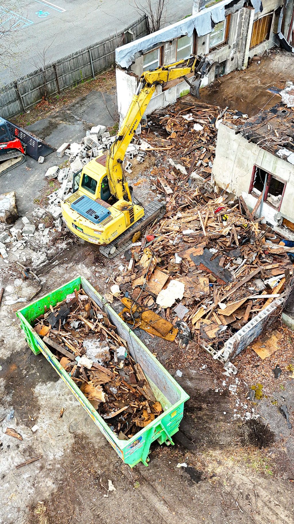 Excavator loading debris into a green dumpster in a demolition site with rubble and a partially ruined building