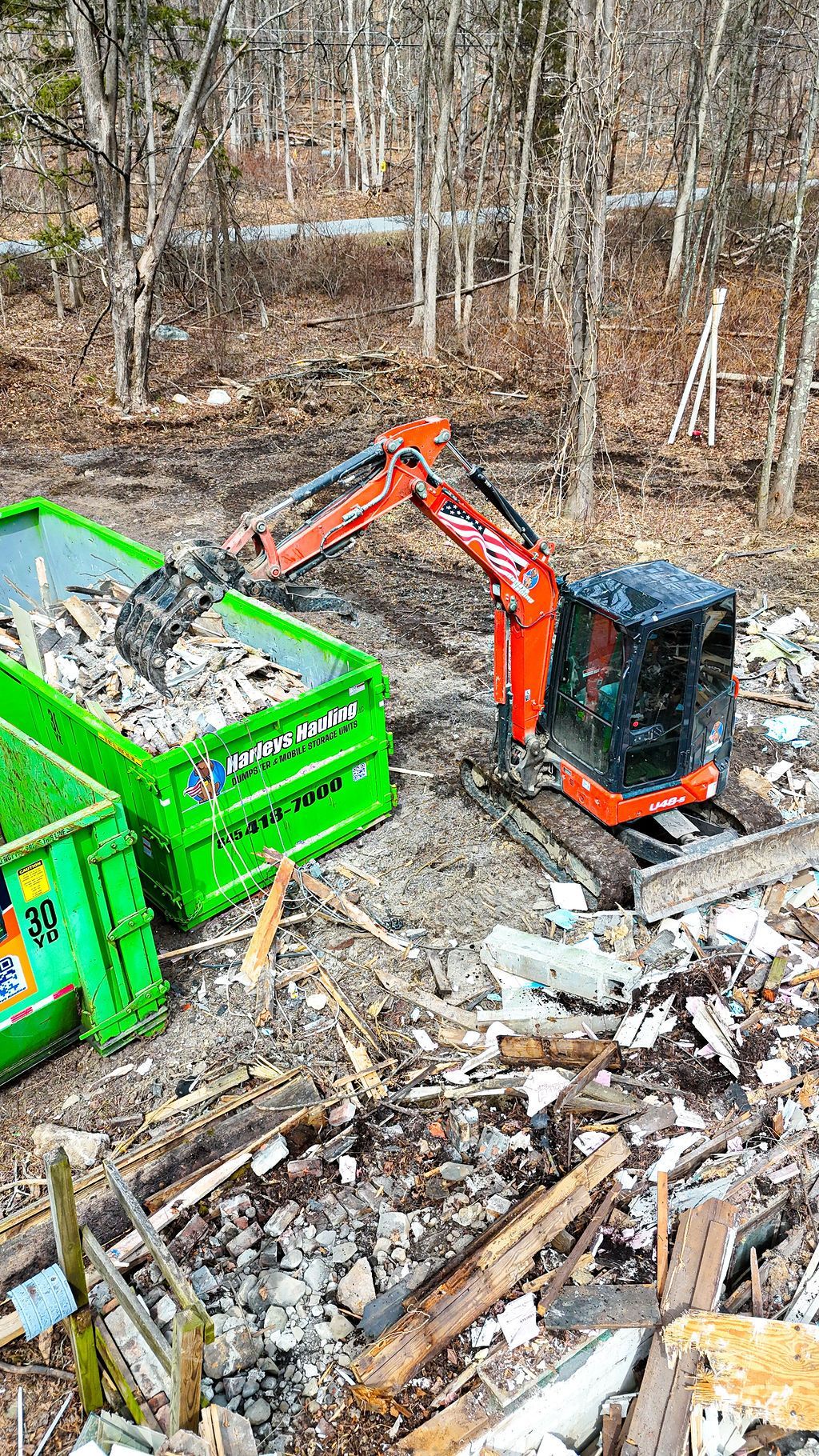 Orange excavator loading debris into a green dumpster in a wooded clearing