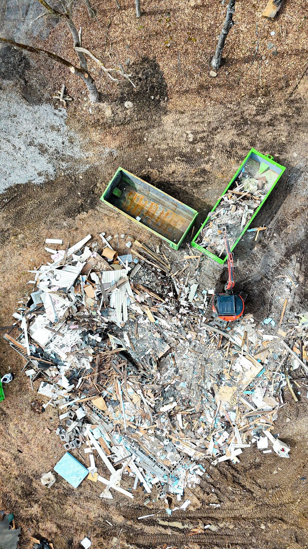 Aerial view of an excavator dumping debris into green dumpsters at a demolition site