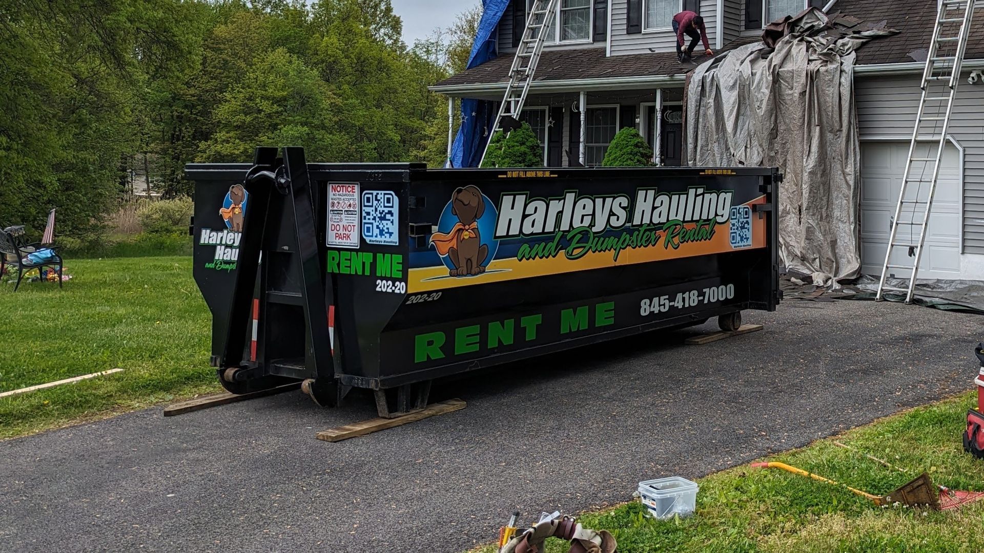 Large black dumpster for Harler’s Hauling and Rent Me parked on a driveway beside a house under repair.