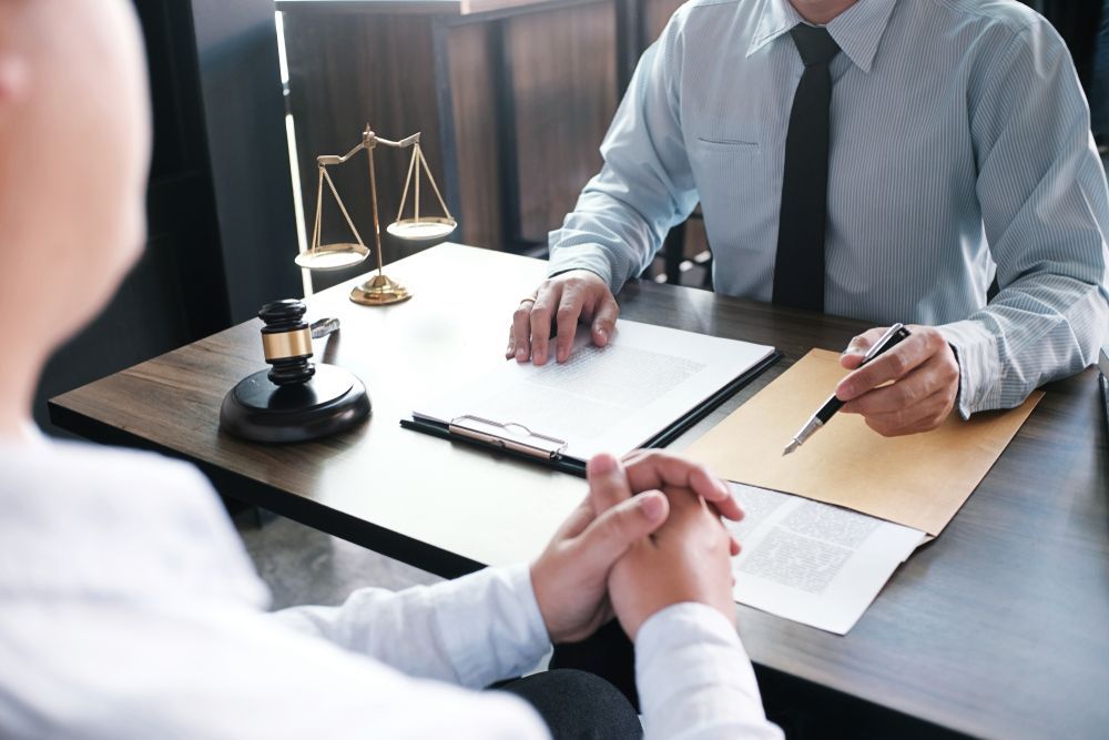 A lawyer and a client sit at a desk with a gavel and scales of justice, discussing legal documents.