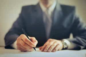 A person in a dark suit sits at a desk, writing on a piece of paper with a pen.