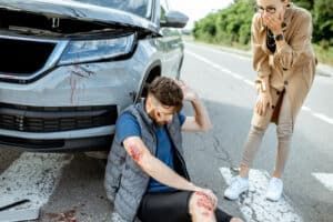 A person sits injured on the road near a damaged car while another person looks on with hands held to their face.