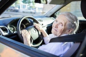 A person sitting in the driver's seat of a car, hands on the steering wheel, looking toward the camera.