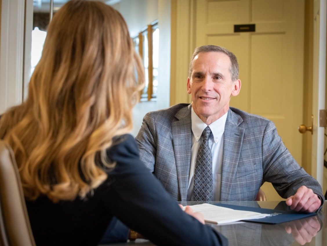 A person in a business suit smiles across a table at an individual in a professional setting during a meeting.