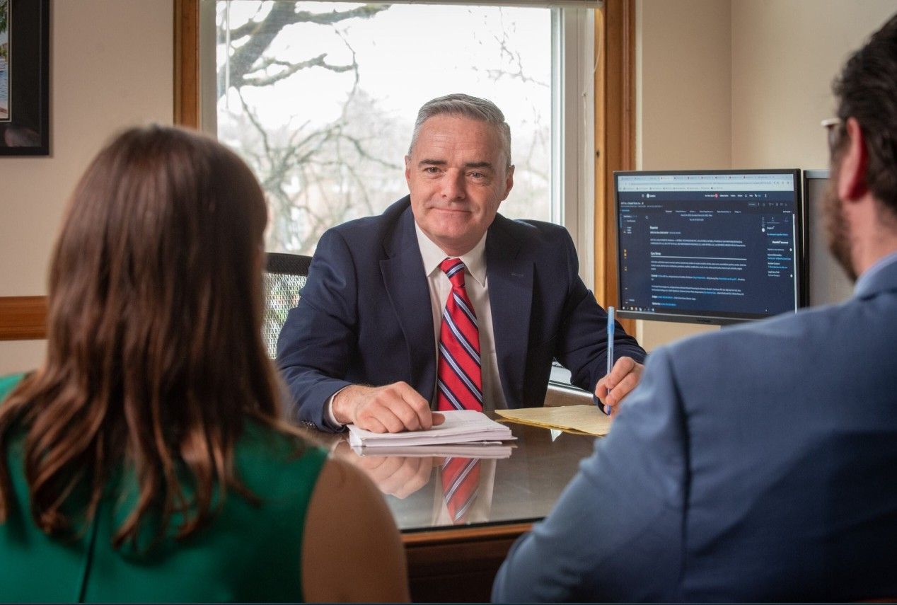 A professional advisor in a suit sits at a desk, facing two people in an office with a computer screen visible behind him.