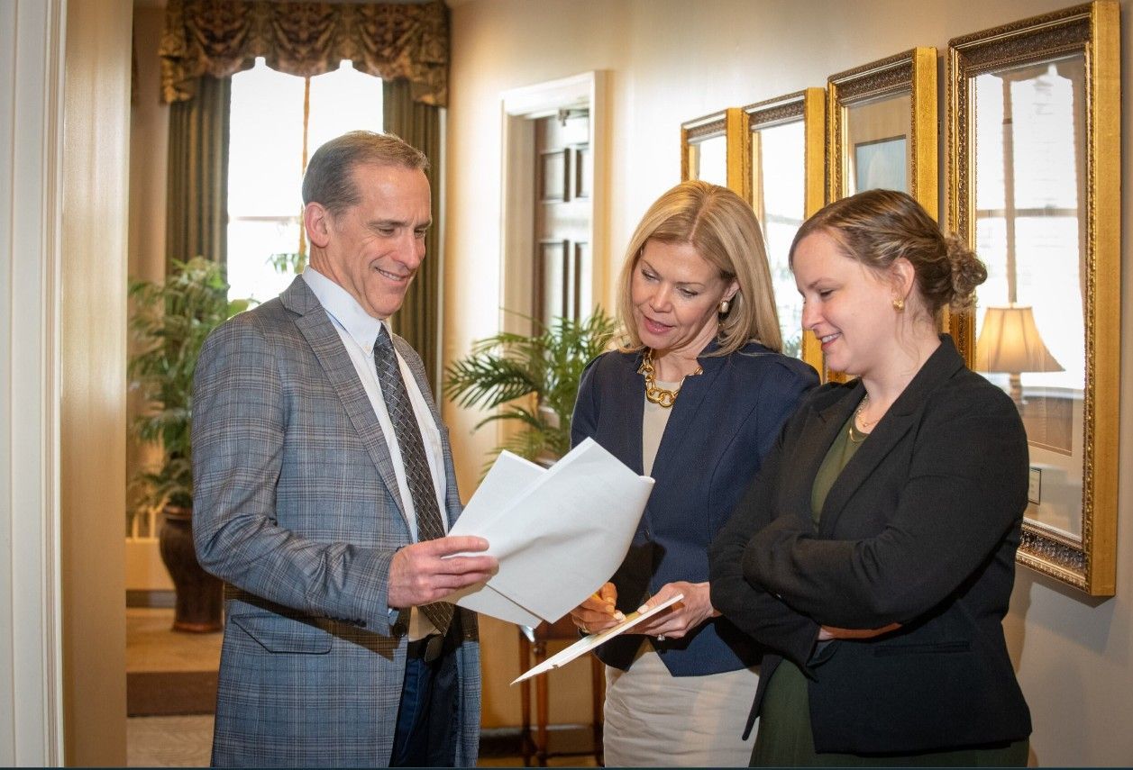 Three professionals stand in a hallway, reviewing documents together while smiling in an office setting.