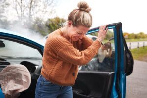A person holding their head in pain after stepping out of a blue car with a deployed airbag and smoke in the background.