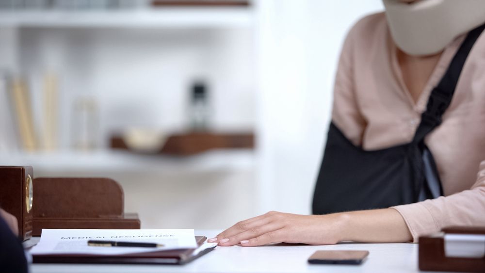A person wearing a neck brace and arm sling sits at a desk with paperwork and a pen.
