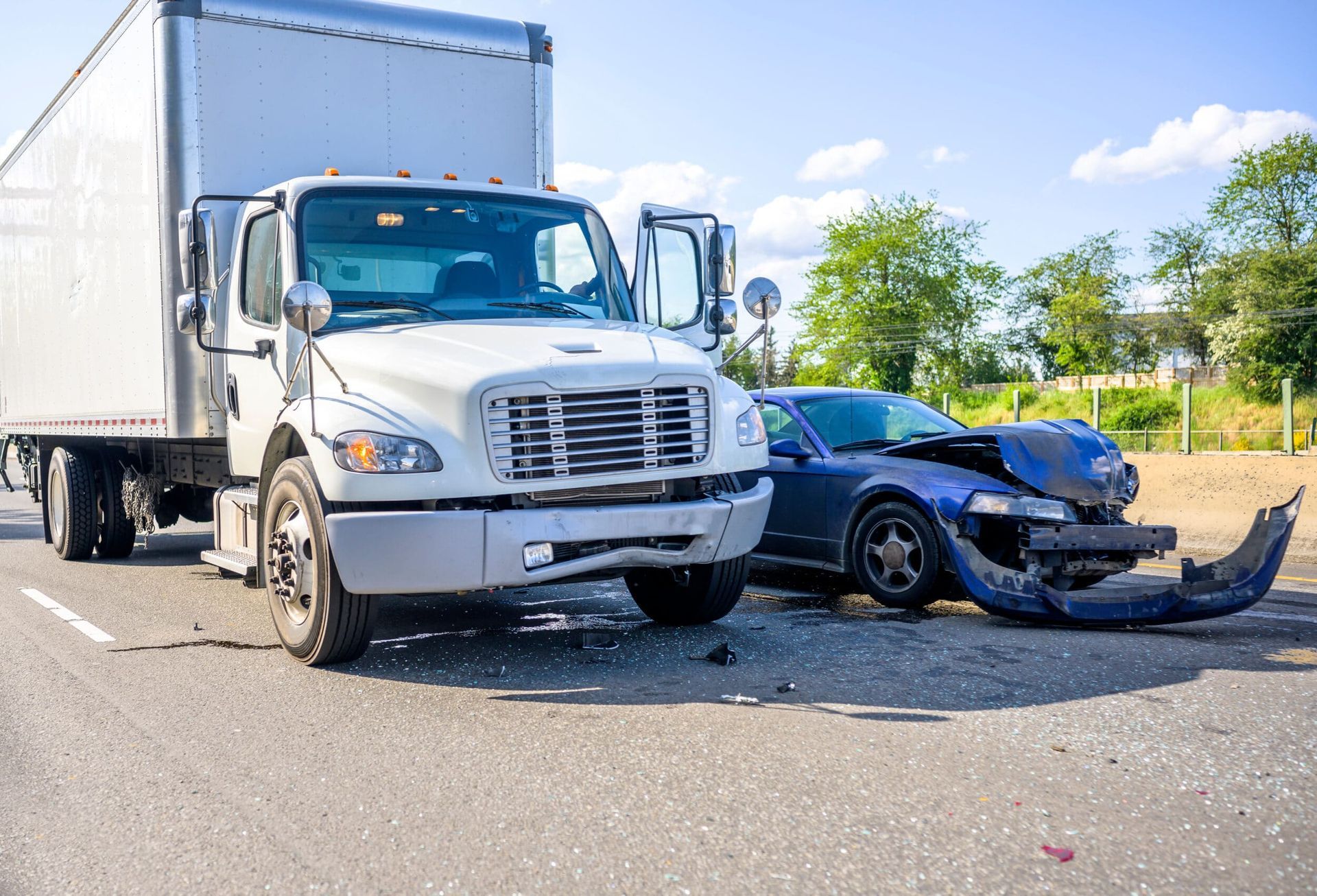 A white semi-truck and a blue car sit on a paved road after a collision, with the car's front bumper hanging off.