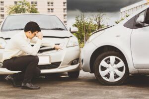 A person crouching in distress while inspecting damage to the front of their car following a collision with another vehicle.