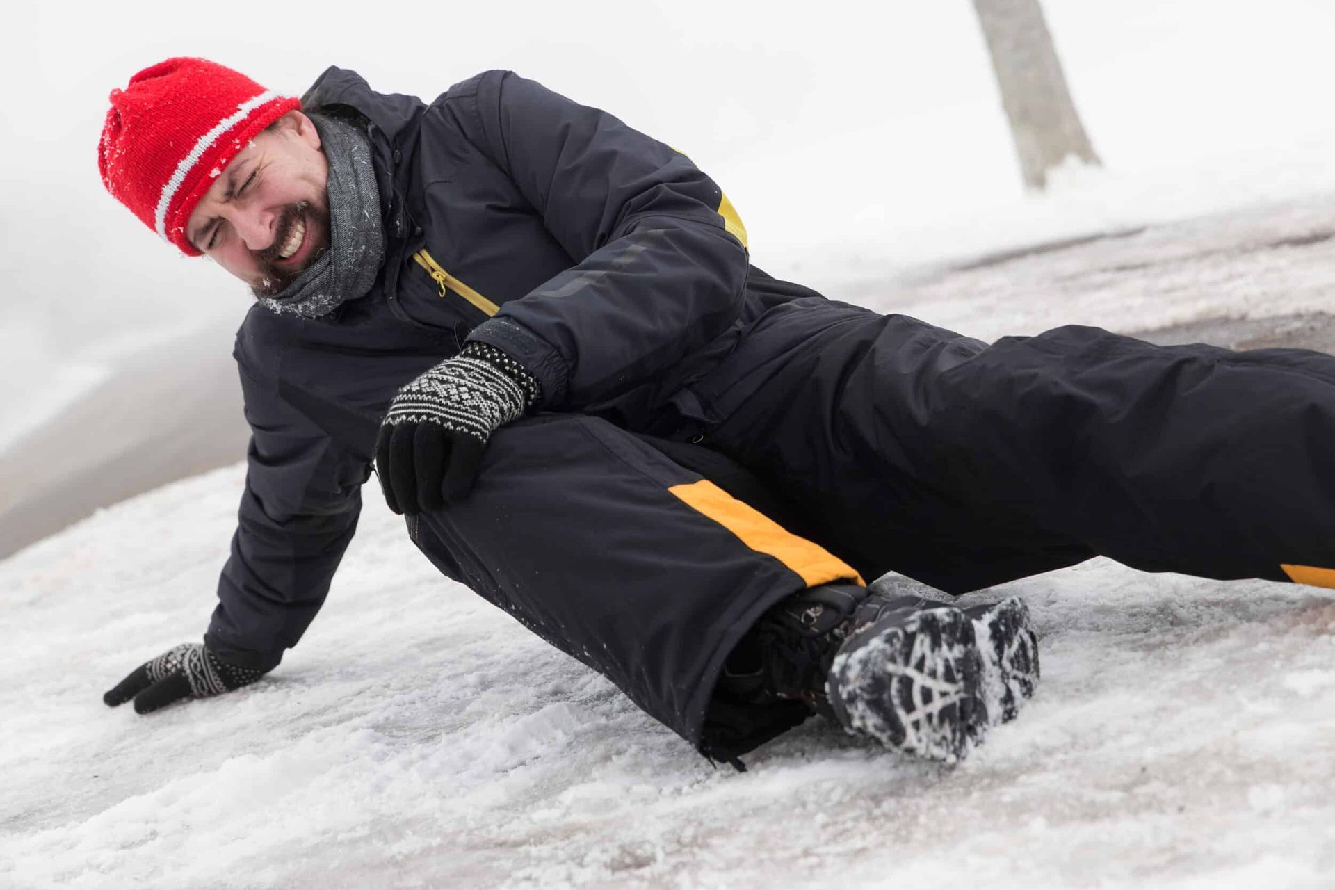 A person in a dark jacket and red hat sits on an icy surface, grimacing in pain after slipping on the snow.