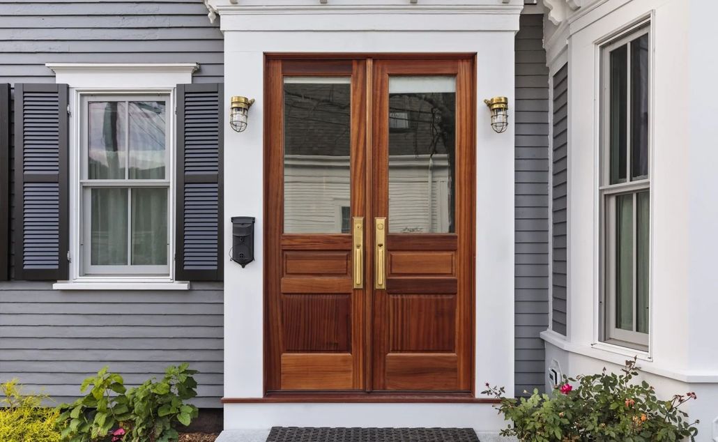 A front entryway with double wooden doors, gray siding, black shutters, and wall-mounted lights on a white frame.