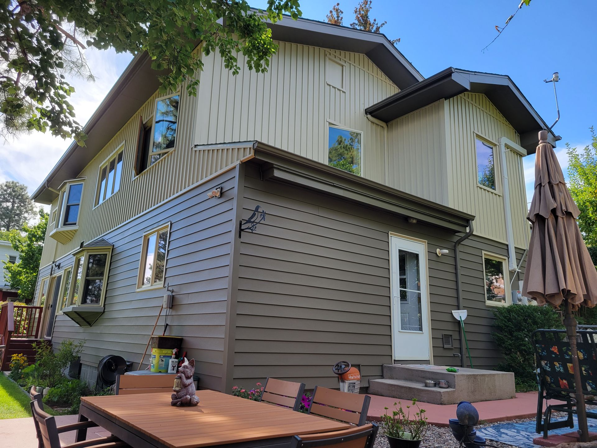 Two-story house with varying tan and brown siding, several windows, and a small porch. An outdoor table and umbrella are visible.