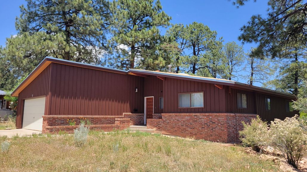 Brown house with a brick facade and a garage, set in a grassy, wooded area under a blue sky.