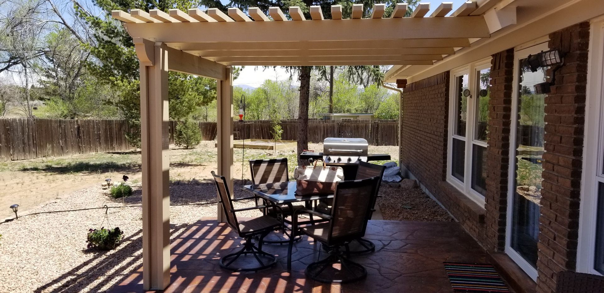 Patio with a table and chairs, a grill, and a pergola. Brick house and fence in the background.