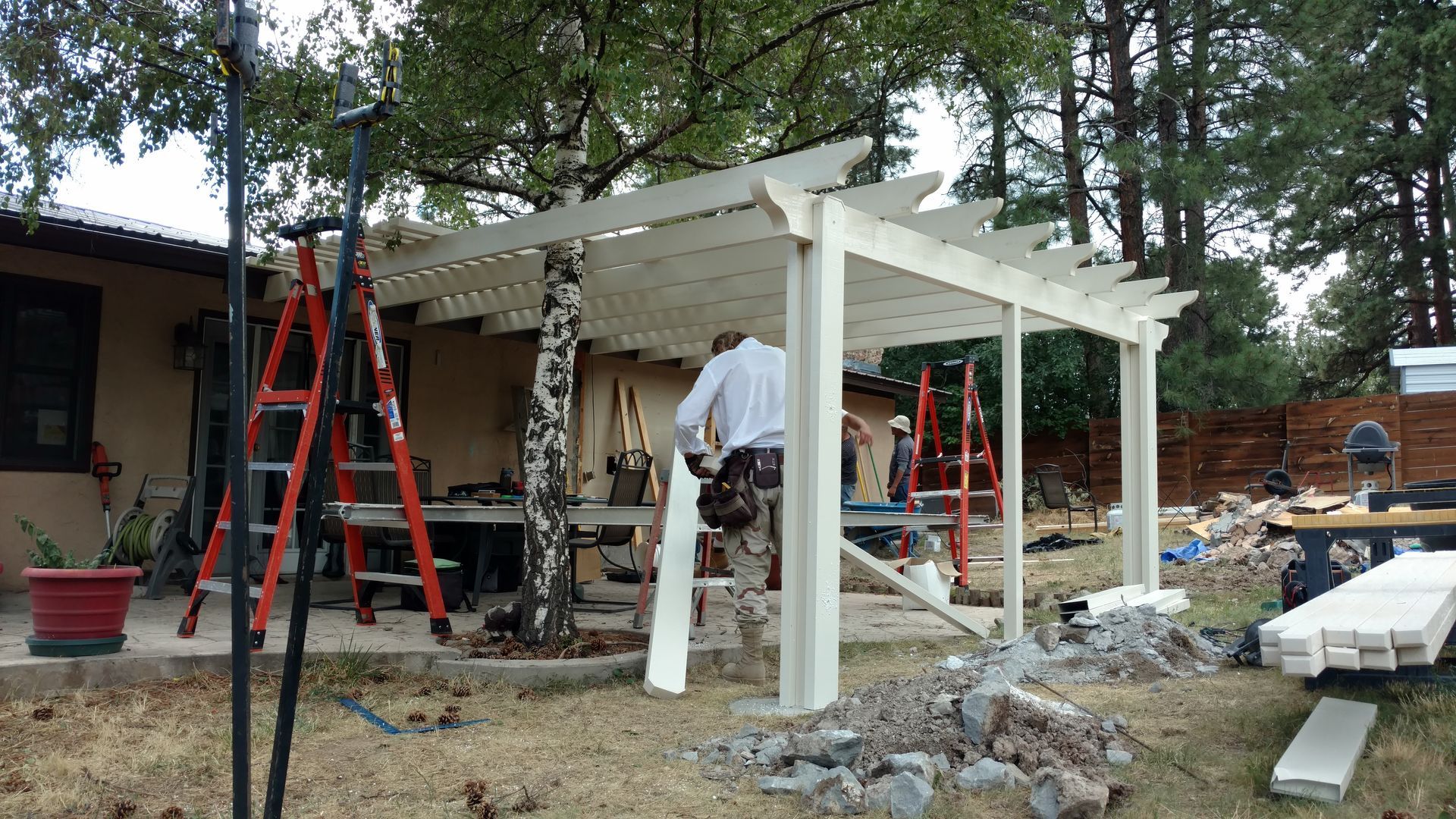 Construction of a white pergola in a backyard; a person works on the structure with ladders nearby.