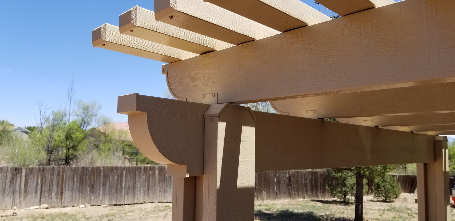 View of a beige pergola with a blue sky background and a wooden fence.