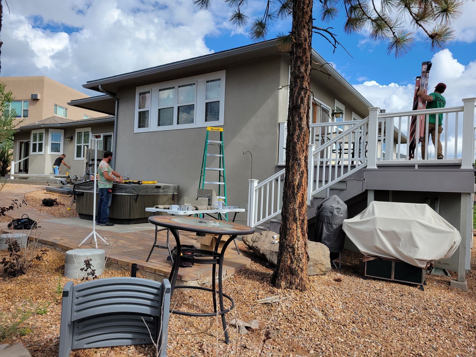 House exterior with workers, hot tub, deck, and trees. Sunny day.