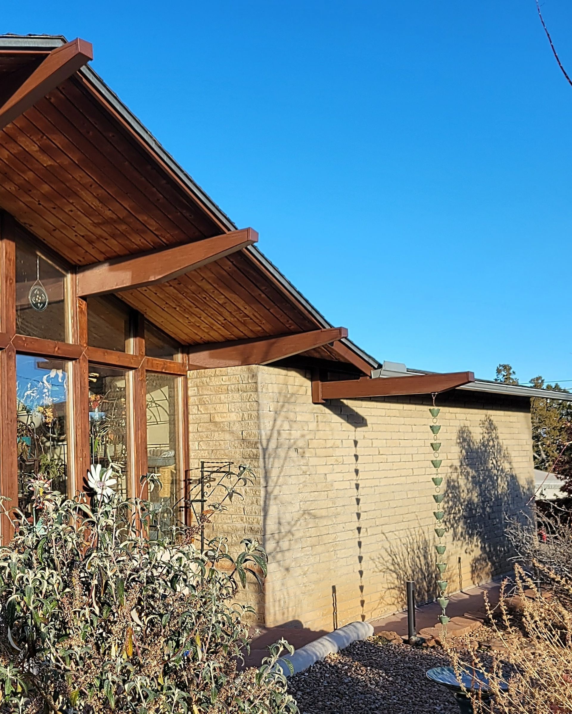 Exterior view of a building with a wood-beam roof, stone wall, and large windows against a clear blue sky.