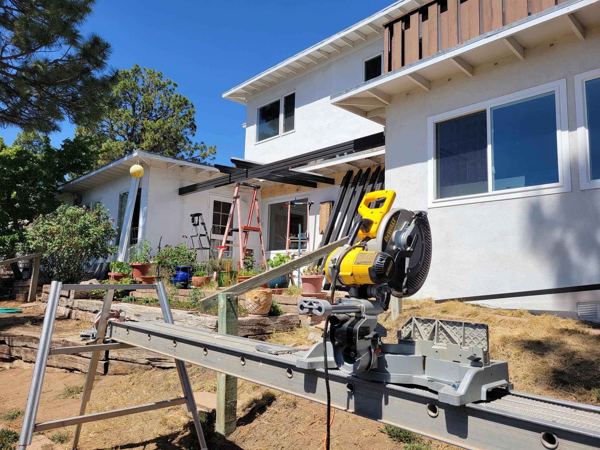 A miter saw on a stand in front of a house, with a ladder and lumber visible, outdoors on a sunny day.