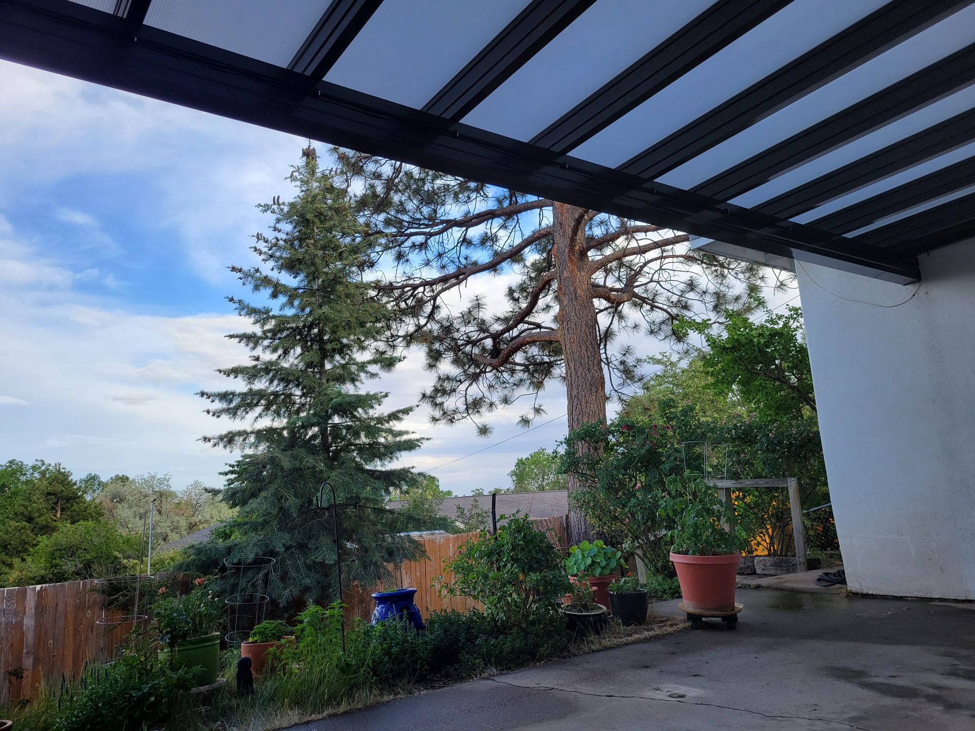 Patio with view of trees, cloudy blue sky. Black awning overhead, garden with potted plants.