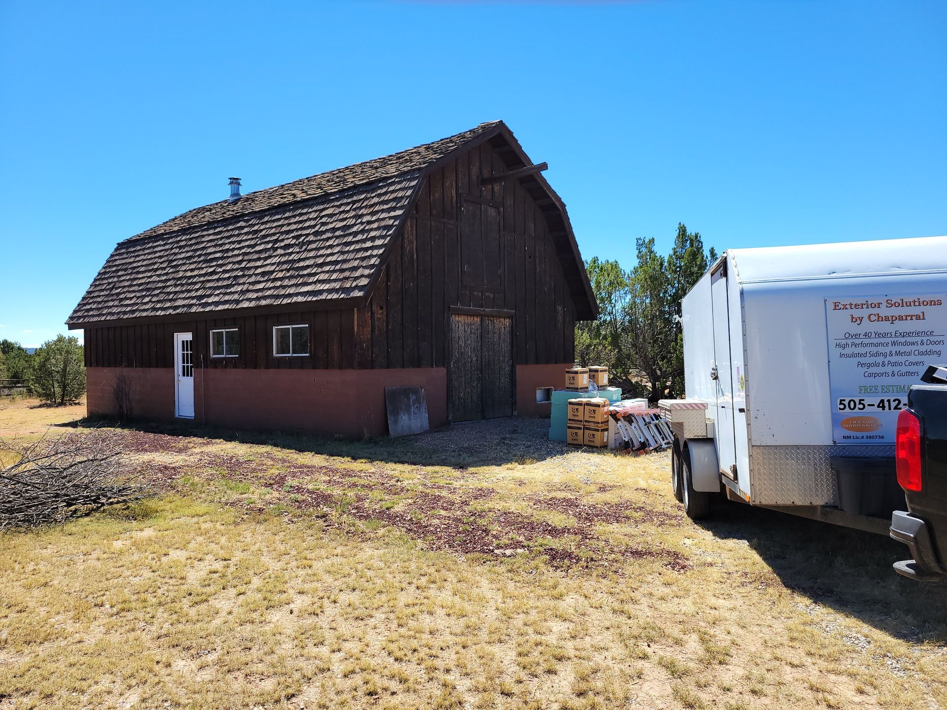 Wooden barn with a dark roof and a small trailer on a sunny day.