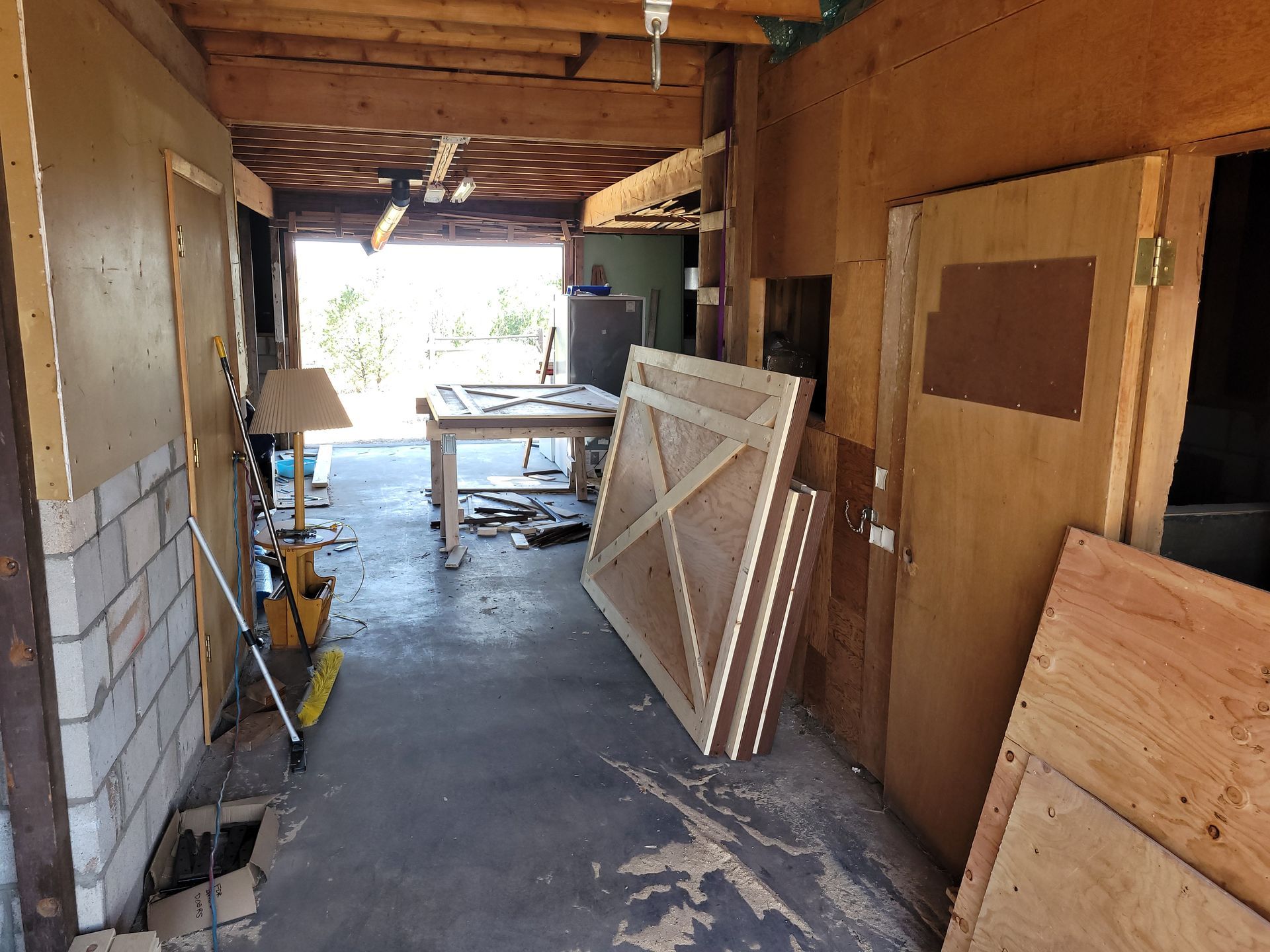 Workshop interior: wood and concrete surfaces, open door to exterior, table with projects, stacked plywood.