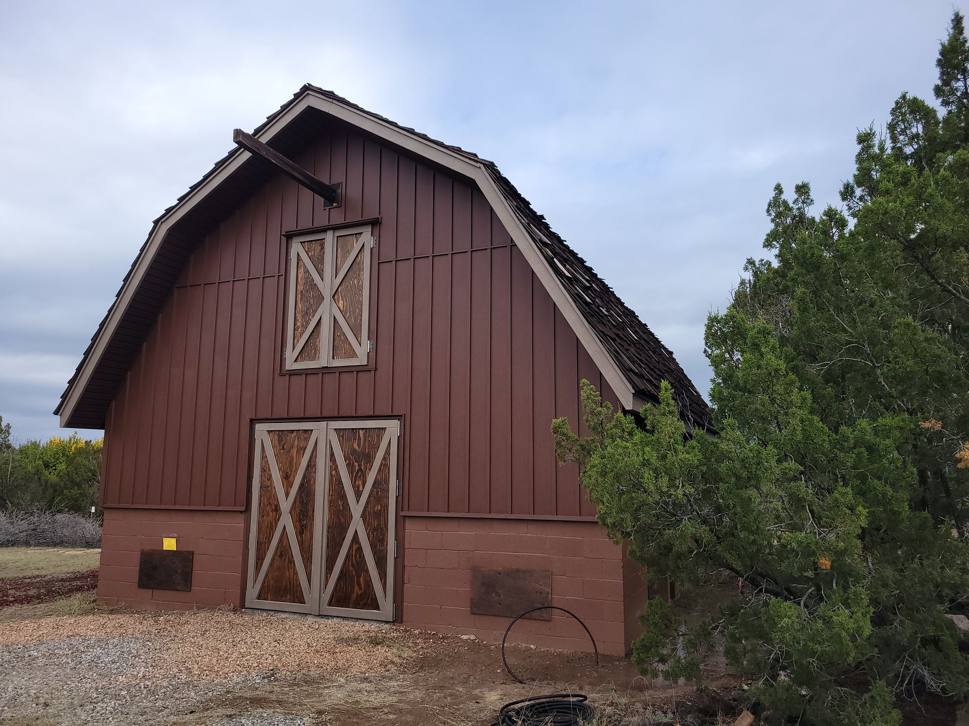 Brown barn with arched roof, wooden doors, and two windows. A cloudy sky is overhead, and a green bush to the right.