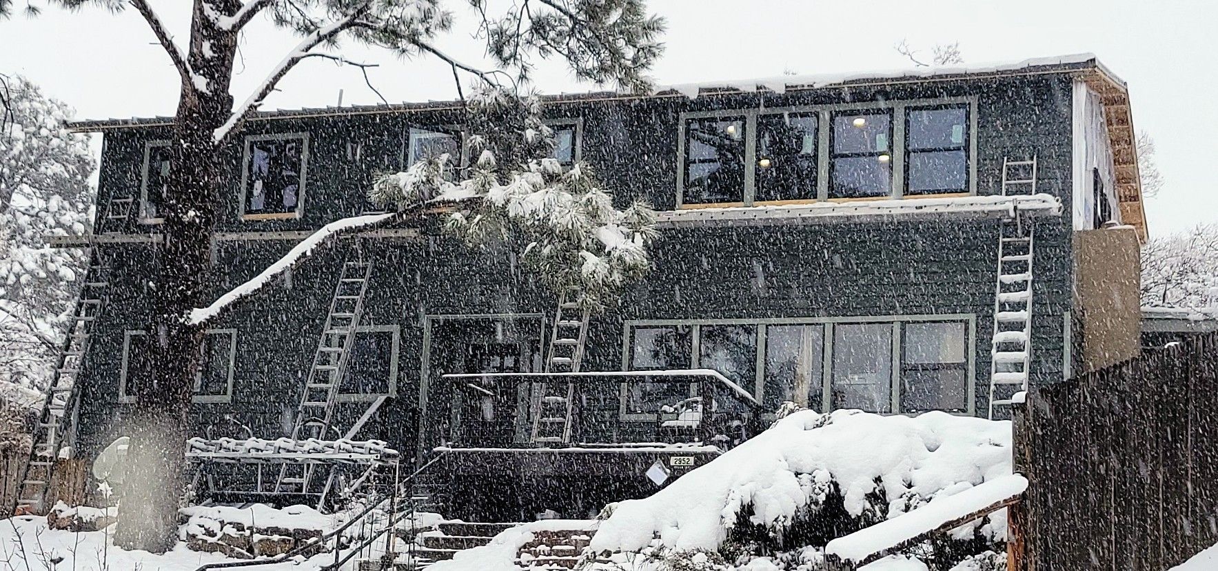 A multi-story house covered in snow during a snowfall, with a large tree in front.
