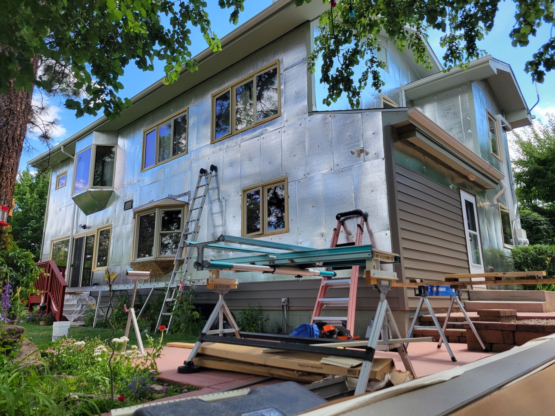 House exterior under construction, covered in silver insulation. Ladders, tools, and lumber present.