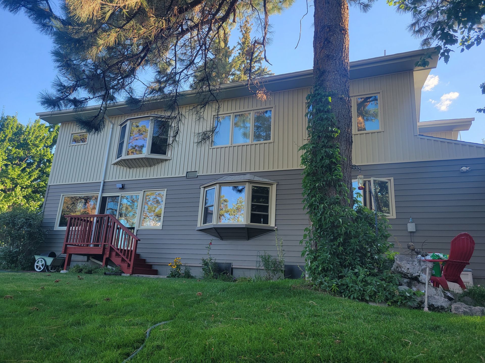 Two-story house with light yellow and gray siding. A tree grows next to the house. Red deck and green lawn.