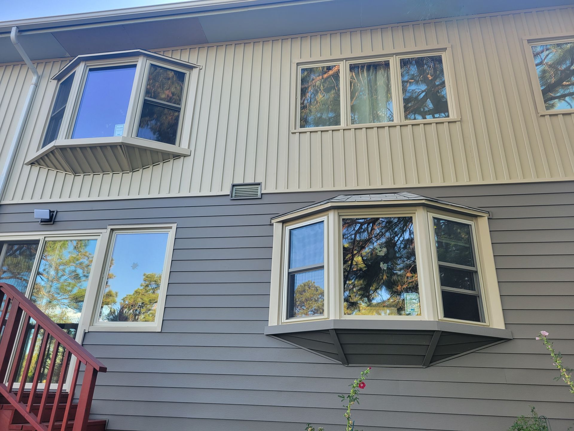 Two-story house exterior with gray and beige siding. Bay windows and other windows are visible.