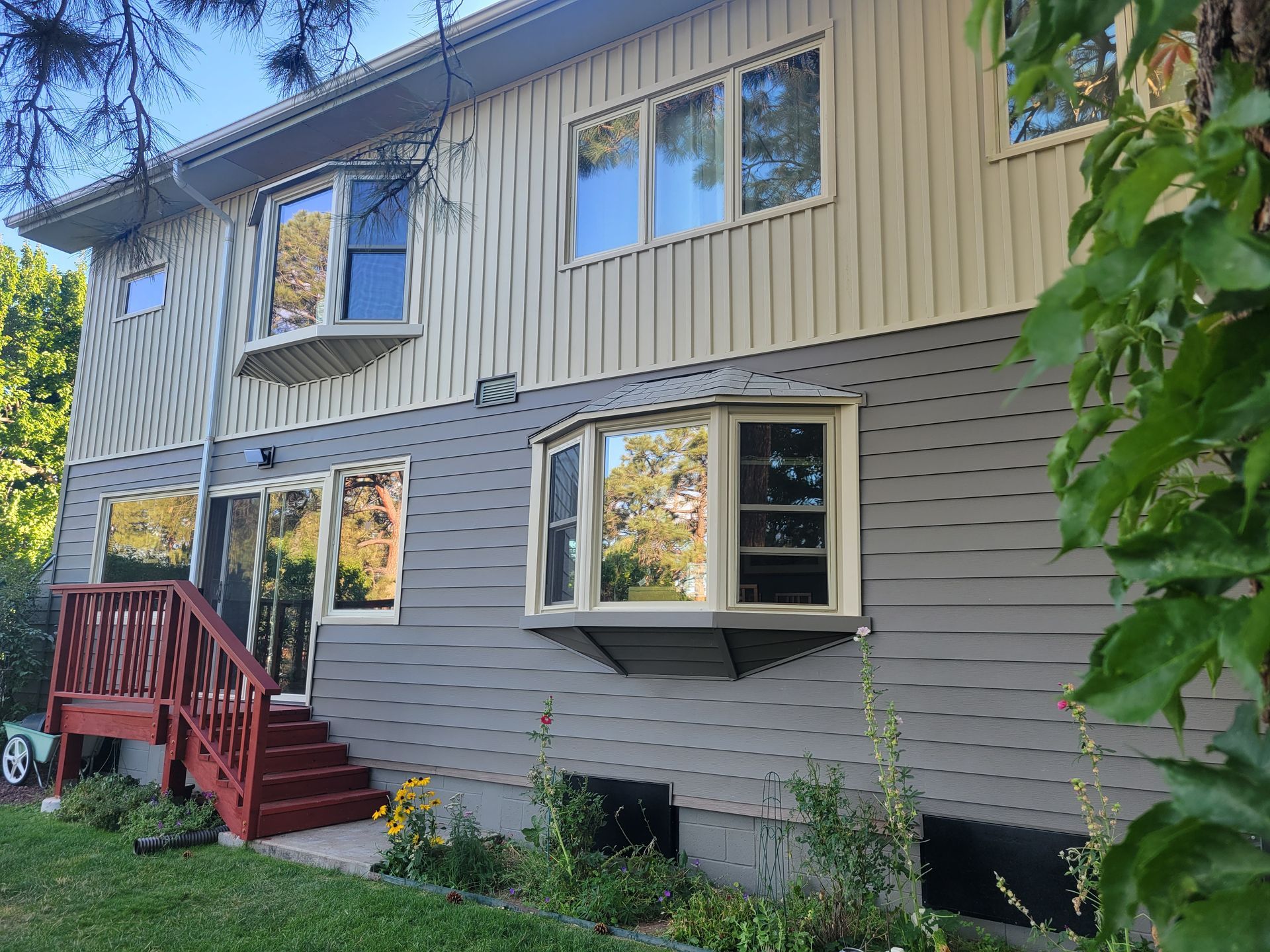 Two-story house with gray and tan siding. Bay windows, sliding glass door, and red stairs. Trees in the background.