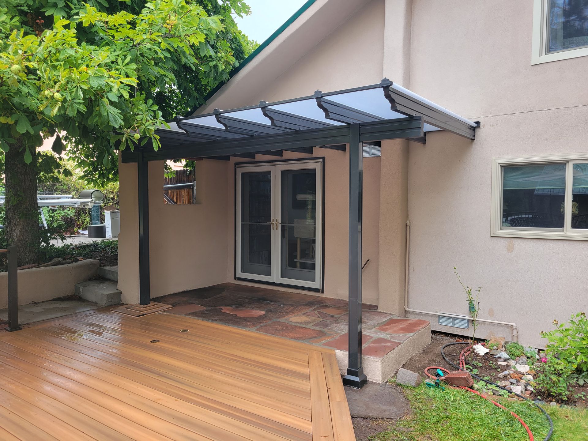 A wooden deck with a black pergola covering a doorway. A tree and house are visible.